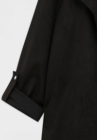 Manche de manteau noir avec le poignet retroussé et maintenu par une patte à bouton-pression, montrant une partie d'une poche avant sur un fond uni.