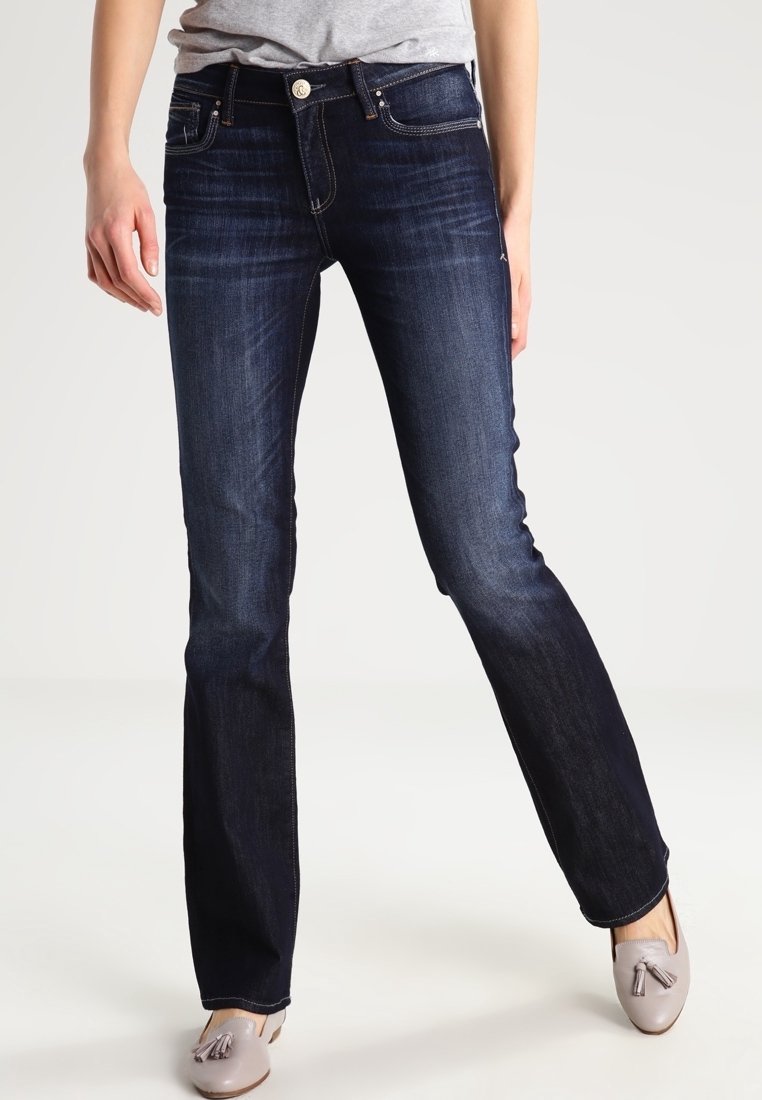 Persoon in donkerblauwe skinny jeans en lichtgrijze tassel loafers, staand met één been iets gebogen.
