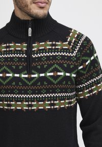 Schwarzer Strickpullover mit hohem Kragen und einem Reißverschluss auf der Vorderseite. Verfügt über grüne, braune und weiße geometrische Muster über der Brust.