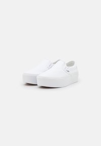 Un paio di sneakers platform bianche senza lacci con suole di gomma spesse, posizionate su uno sfondo chiaro e semplice.