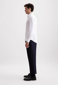 Camicia bianca con bottoni e maniche lunghe, abbinata a pantaloni blu scuro e scarpe nere. Tessuto liscio e vestibilità su misura.