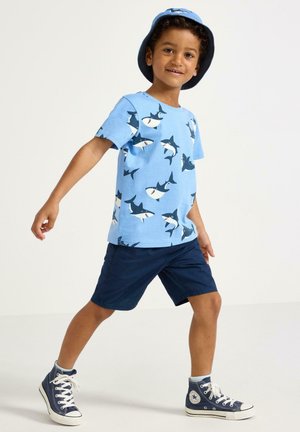 Nuori poika, jolla on sininen haukkuva t-paita, siniset shortsit, sopiva haukkupainatushattu ja tummansininen korkeavartiset tennarit, kävelee eteenpäin.