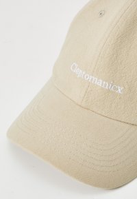 Gorra de algodón beige con una superficie texturizada; cuenta con el texto bordado en blanco "Cleptomanicx" en la parte frontal; visor curvado y detalles cosidos.