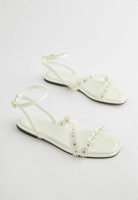 Weiße flache Sandalen mit quadratischer Zehenpartie und offenem Design. Mit dekorativen Gänseblümchen-Akzenten entlang der Riemen und einem dünnen Knöchelriemen mit Schnalle.