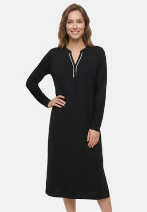 Vrouw die een zwart midi-jurk met lange mouwen draagt, voorzien van een V-hals met knopen en witte bies, glimlachend met een hand op haar heup.