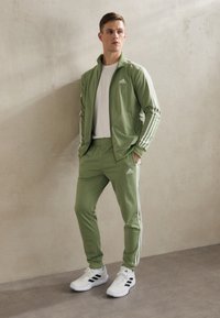 Zeleni sportski trenirke s bijelim prugama, jakna na patentni zatvarač i hlače s elastičnim pojasom. Nošeno s bijelom majicom i tenisicama s crnim naglascima.