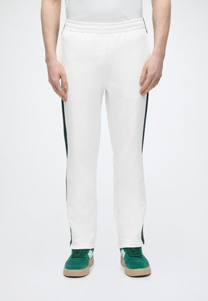 Persona che indossa pantaloni sportivi bianchi con strisce laterali scure e sneakers in camoscio verde, in piedi davanti a uno sfondo semplice.