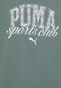 Grønn bomull t-skjorte med stor hvit slitt tekst "PUMA sports club" og en liten hvit puma-logo nederst til høyre.