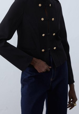 Persona con una chaqueta negra ajustada con botones dorados y jeans azules de cintura alta, con la mano apoyada cerca de la cintura.