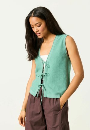 Woman wearing a sleeveless mint green knit vest with tie closures over a white top and loose brown pants, looking downward with one hand in pocket.