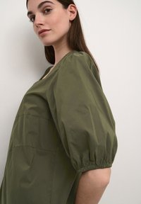 Olive zaļš pārāk liels kleita ar plašu dekoltē un pūkainām īsām piedurknēm, ar savelkošiem elastīgiem aprocēm un gludu, matētu auduma tekstūru.