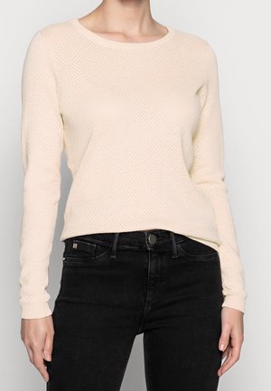 Maglione lavorato a maglia di un beige chiaro con scollatura tonda, maniche lunghe e motivo testurizzato. Abbinato a jeans neri con una silhouette aderente.