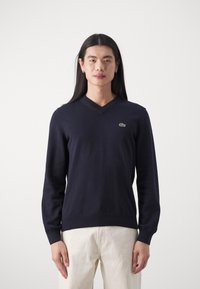 Námornícky modrý sveter s výstrihom do V a dlhými rukávmi, vyrobený z pleteného materiálu. Má malú zelenú nášivku Lacoste na ľavom hrudníku.