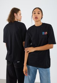 Black cotton t-shirts feature a relaxed fit, short sleeves, and colorful logo patches on the front. Denim pants are worn with open hems.