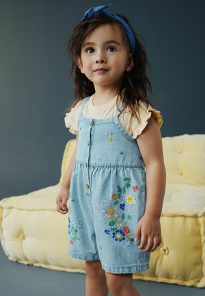 Fată tânără cu păr lung și închis la culoare, purtând o bentită albastră, o bluză galbenă cu mâneci volanate și o salopetă din denim cu broderii florale colorate, stând în interior.