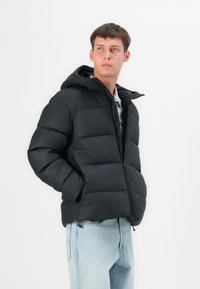 Melnā pūkainā jaka ar kapuci, ar rāvējslēdzēju un quilted rakstu. Iebūvēti elastīgi piedurkņu gali un sānu kabatas, valkāta virs gaiši ziliem džinsiem.