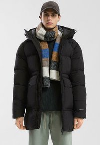 Čierna prešívaná bunda s kapucňou, s doskou na zips a dvoma prednými vreckami. Nosí sa s viacfarebným prúžkovaným šálom a zeleným svetrom.