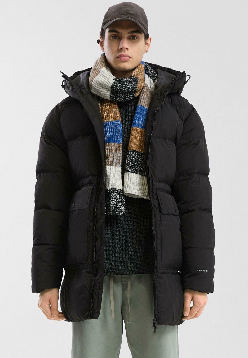 Čierna prešívaná bunda s kapucňou, s doskou na zips a dvoma prednými vreckami. Nosí sa s viacfarebným prúžkovaným šálom a zeleným svetrom.