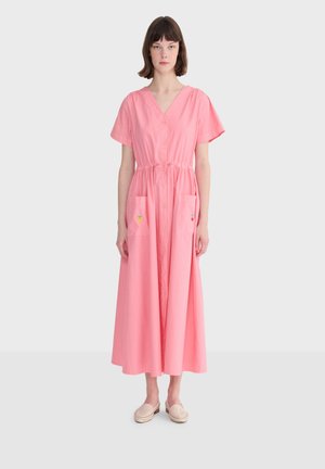 Kobieta ubrana w różową, midi sukienkę na guziki z krótkim rękawem, z troczkiem w pasie i przednimi kieszeniami, stojąca na jasnoszarym tle.