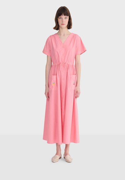 Femme portant une robe midi rose à manches courtes, boutonnée sur le devant, avec taille à cordon et poches devant, debout sur un fond gris clair.