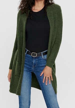 Kvinde iført en mørkegrøn strikket cardigan, sort top, blå jeans og sort bælte, med ringe på sin højre hånd, der hviler på låret.
