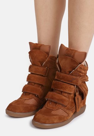 Baskets compensées montantes en daim marron avec trois sangles velcro, portées sur des jambes nues, sur fond blanc.