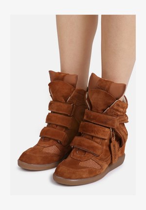 Braune High-Top-Sneaker aus Wildleder mit Keilabsatz und drei Klettverschlüssen, getragen an nackten Beinen vor weißem Hintergrund.