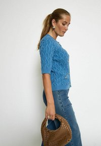 Ljusblå stickad cardigan med ett texturerat kabelmönster, korta ärmar och guldknappar. Matcher med högmidjade denimjeans och en brun vävd väska.