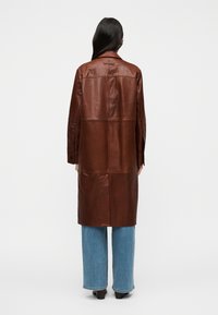 Καφέ δερμάτινο μακρύ παλτό με ίσια γραμμή, που διαθέτει πίσω κόψιμο και μανίκια με λεπτομέρεια κουμπιών, φορεμένο πάνω από ανοιχτό μπλε τζιν παντελόνα με φαρδιά μπατζάκια.