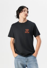 T-shirt preta de algodão com mangas curtas, apresentando uma pequena impressão laranja "RIDE THE RAILS" no lado esquerdo do peito. Usada com jeans de tom azul claro.