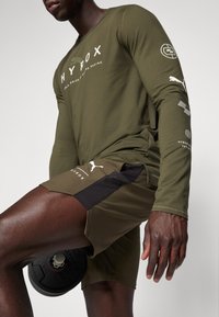 Olijfgroene longsleeve shirt met "HYROX" logo en zwarte accenten; gecombineerd met bijpassende shorts met een zwart paneel en wit logo, terwijl hij een gewicht vasthoudt.