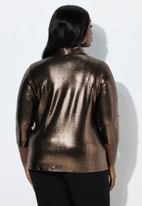 Bronsefärgad metallisk långärmad topp med ett texturerat mönster, med krage och rak skärning, tillsammans med svarta byxor.