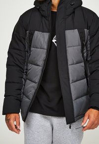 Padded jacket w czarno-szarym kolorze, z zamek błyskawicznym z przodu, dwoma bocznymi kieszeniami i kapturem. Materiał wydaje się wodoodporny i ma pikowaną strukturę.