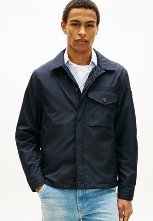 Ung mand iført navyblå let jakke over hvid skjorte og lyseblå jeans, med hænderne i lommerne, vendt fremad.