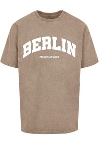 Hnědý tričko s krátkým rukávem, na kterém je v tučném bílém obloukovém písmu napsáno "BERLÍN" a pod tím "MERCH●DE", s jemnou, mírně vybledlou texturou.