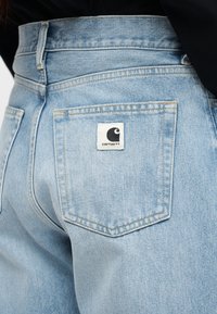 Ανοιχτό μπλε τζιν παντελόνι που φοριέται από άτομο, με έμφαση στην πίσω τσέπη όπου φαίνεται το λευκό λογότυπο Carhartt.