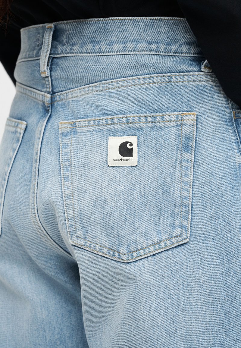 Lichtblauwe spijkerbroek gedragen door persoon, met focus op achterzak met wit Carhartt-logoplaatje.