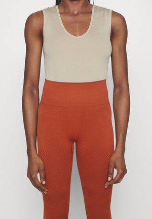 Person iført en ærmeløs beige top og højtaljede rustfarvede teksturerede leggings, stående mod en ensfarvet baggrund.