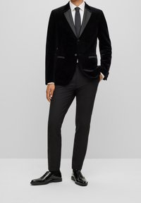 Veste de smoking en velours noir avec revers en satin, associée à un pantalon noir et des chaussures brillantes. Présente une coupe slim et des éléments de design minimalistes.
