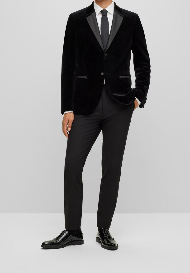 Veste de smoking en velours noir avec revers en satin, associée à un pantalon noir et des chaussures brillantes. Présente une coupe slim et des éléments de design minimalistes.
