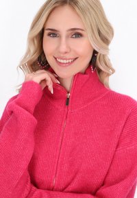Maglione fucsia con zip, texture a coste, colletto alto e dettaglio della zip dorata. Il modello tiene la mano vicino al viso, mettendo in risalto il design.