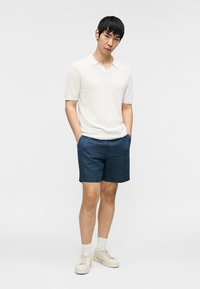 Homme debout les mains dans les poches, portant un polo blanc à manches courtes, un short bleu marine, des chaussettes blanches et des baskets crème.