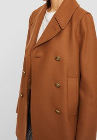Bruine dubbelgeknoopte jas van gestructureerde stof. Heeft een grote kraag, gouden knopen en zijzakken met een getailleerde pasvorm.