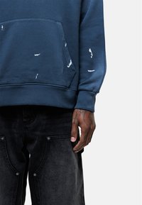 Felpa blu con tasca frontale, caratterizzata da segni di usura bianchi. Abbinata a jeans scuri, che mostrano dettagli in metallo e texture.