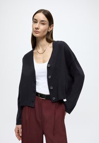 Cardigã preto de malha com corte solto, decote redondo e cinco botões. Combinado com uma camisola branca de alças e calças de cintura alta em bordô.