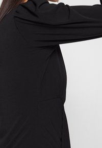 Svart klänning med långa ärmar med gathered-detaljer, en figursydd midja och ett mjukt, texturerat tyg. Sid sömsdesign som synligt kurvar.