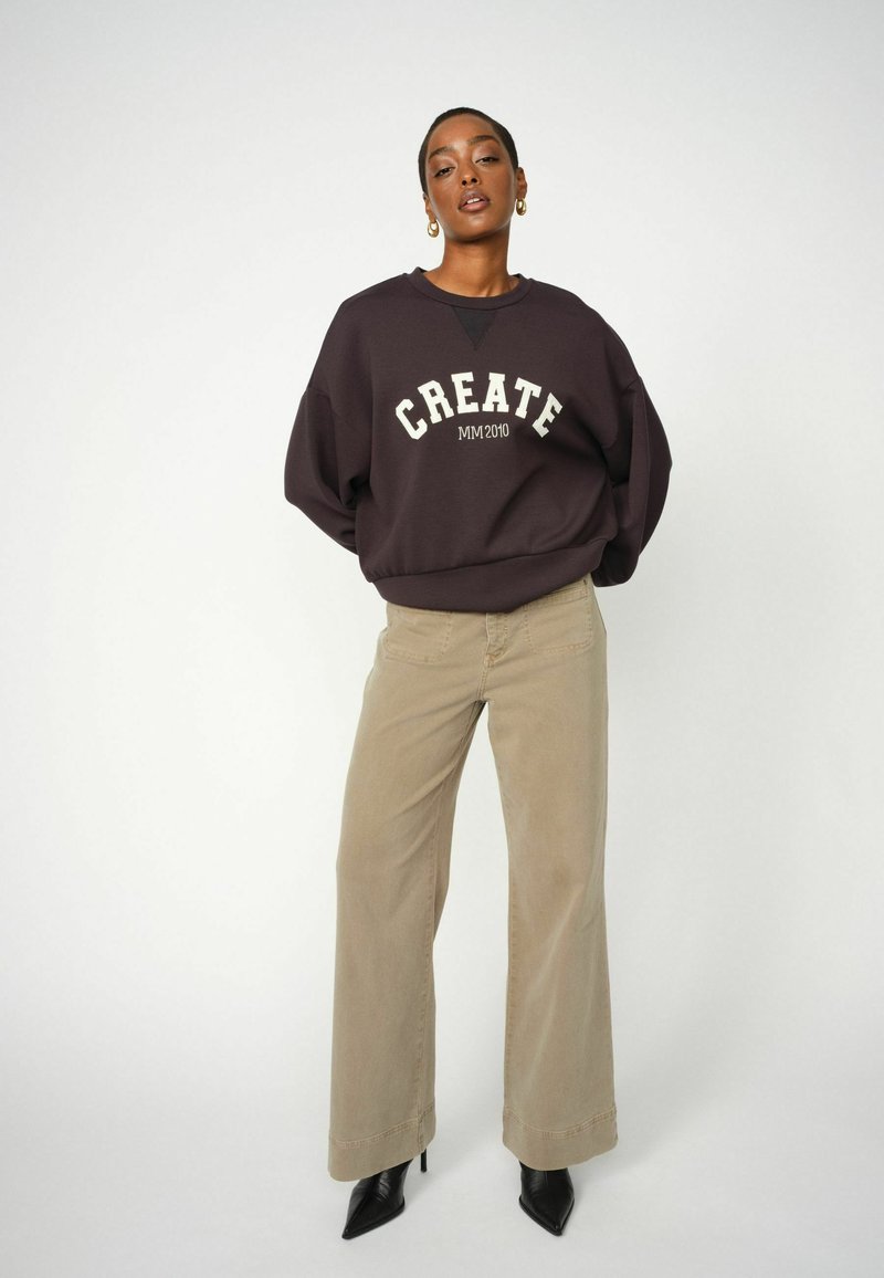 Sweatshirt violet foncé avec impression blanche "CREATE MM2010", associé à un pantalon large beige et des escarpins noirs pointus. Tissu lisse, coupe décontractée.