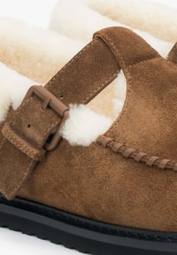 Brązowe zamszowe kapcie z miękkim białym wyściełaniem, regulowanym paskiem z klamrą i teksturowaną podeszwą. Szczegółowe przeszycia wzdłuż krawędzi.