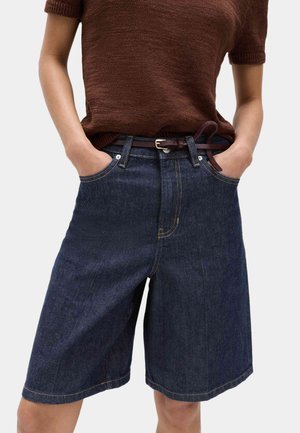 Person iført mørke denim knælange shorts med et mørkebrunt bælte og en kortærmet brun striktrøje, hænderne i lommerne.