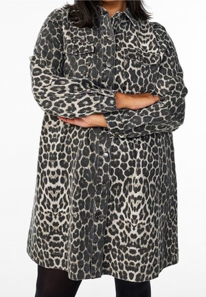 Person iført en knaplukket, langærmet leopardprint jakke med armene krydset, stående mod en ensfarvet hvid baggrund.